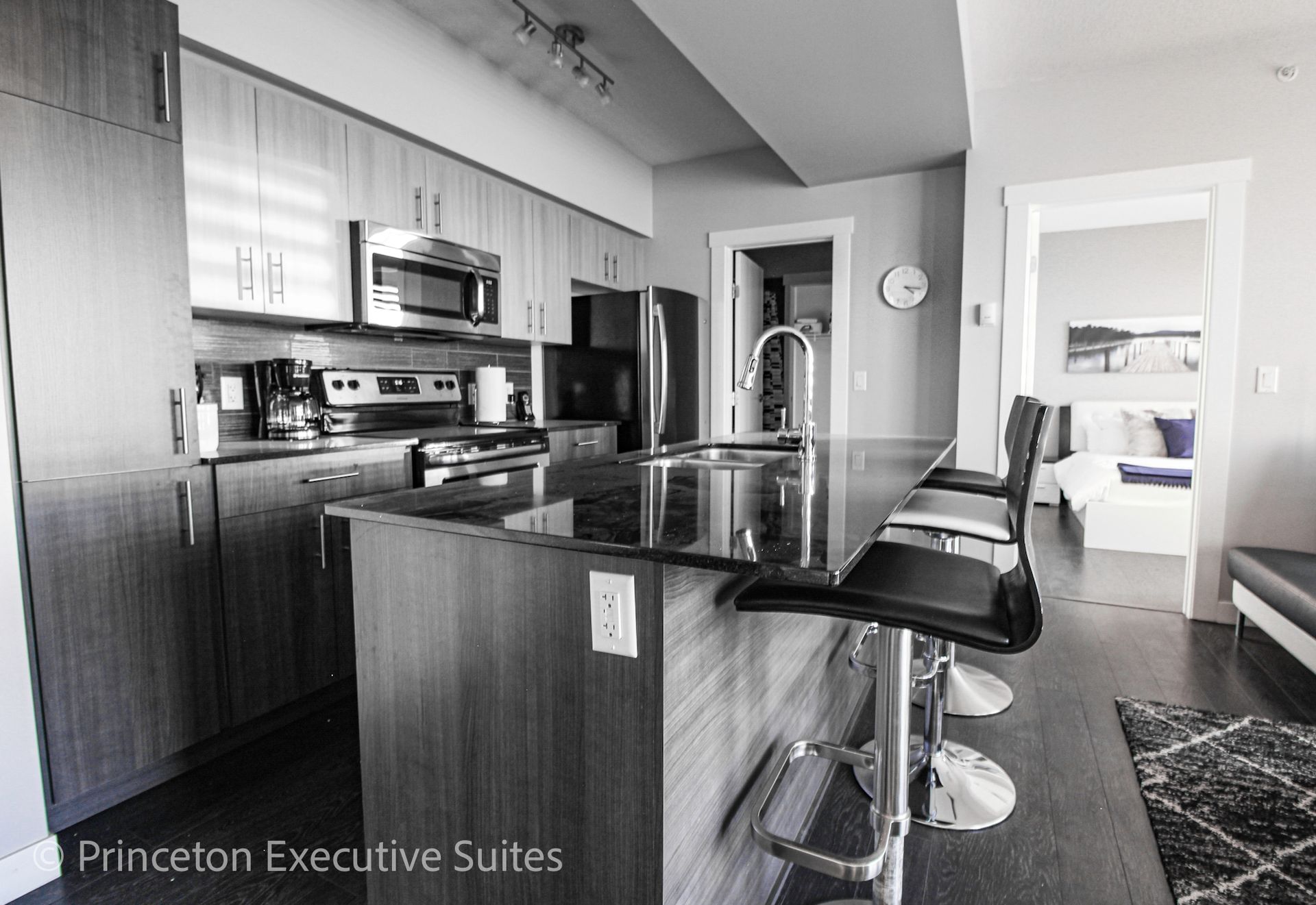 Modern kitchen with island and bar stools; view into bedroom. Black and white.