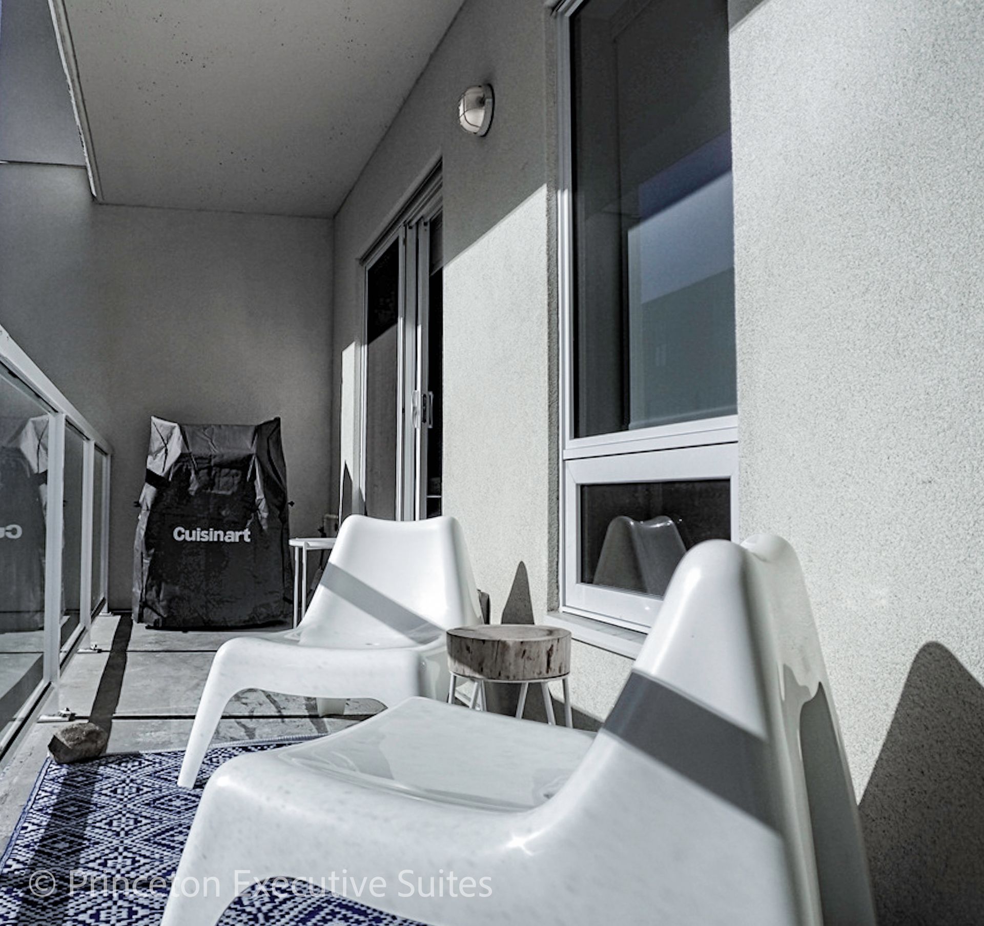 Balcony with white chairs, rug, bag, and a small table next to sliding glass door.