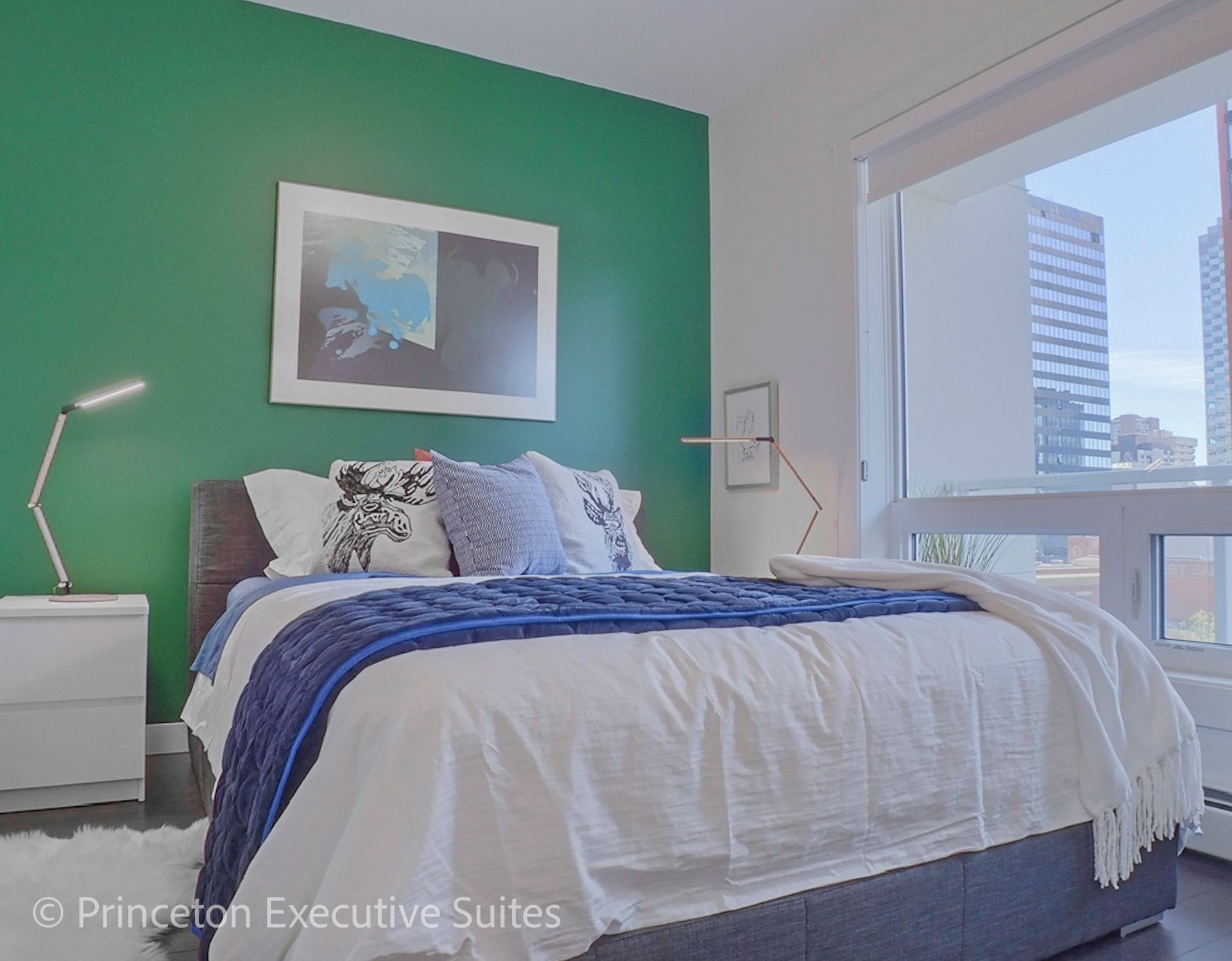 Bedroom with green accent wall, bed with blue blanket, two lamps, and city view.
