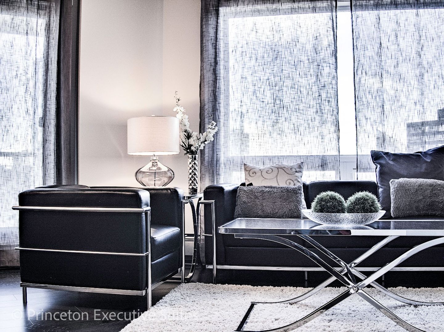 A modern furnished apartment with black leather couch chair and chair with metal and glass coffee table in front of it.