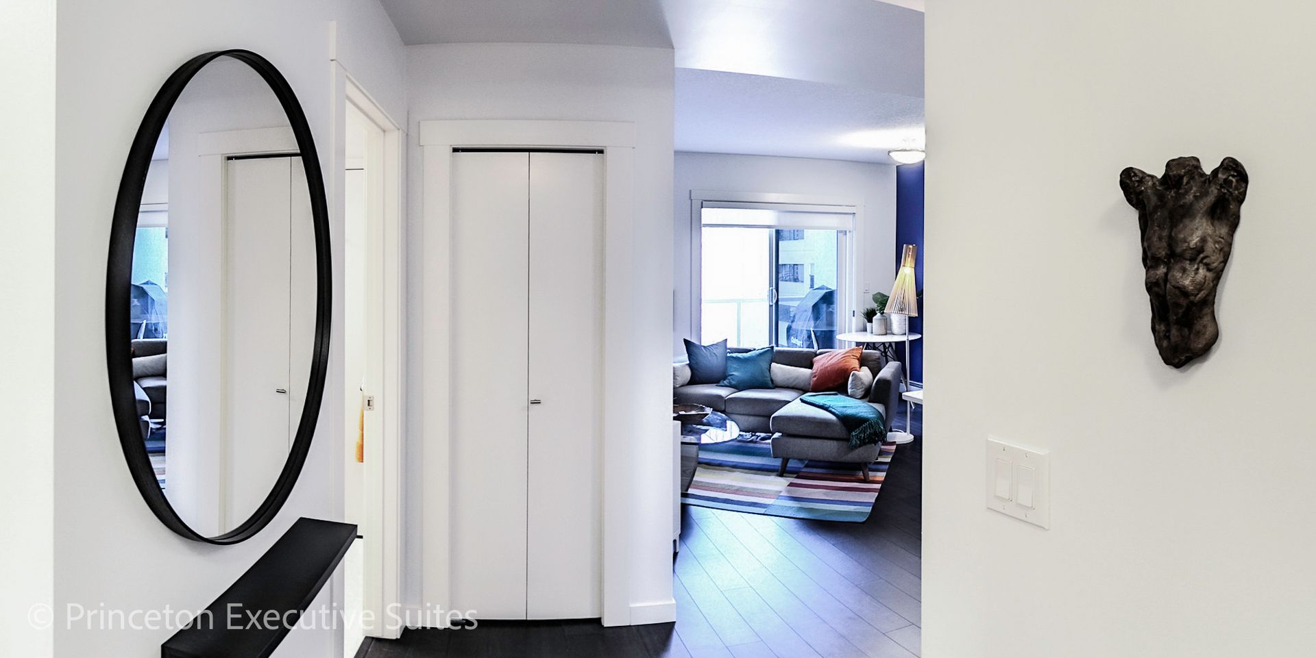 Hallway with oval mirror, white doors, and wall art leading to a living room with a sofa and rug.