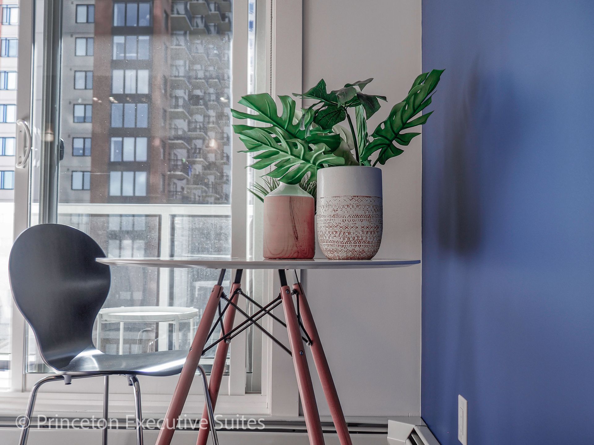 A small table with plants and a chair near a window with a city view, against a blue wall.