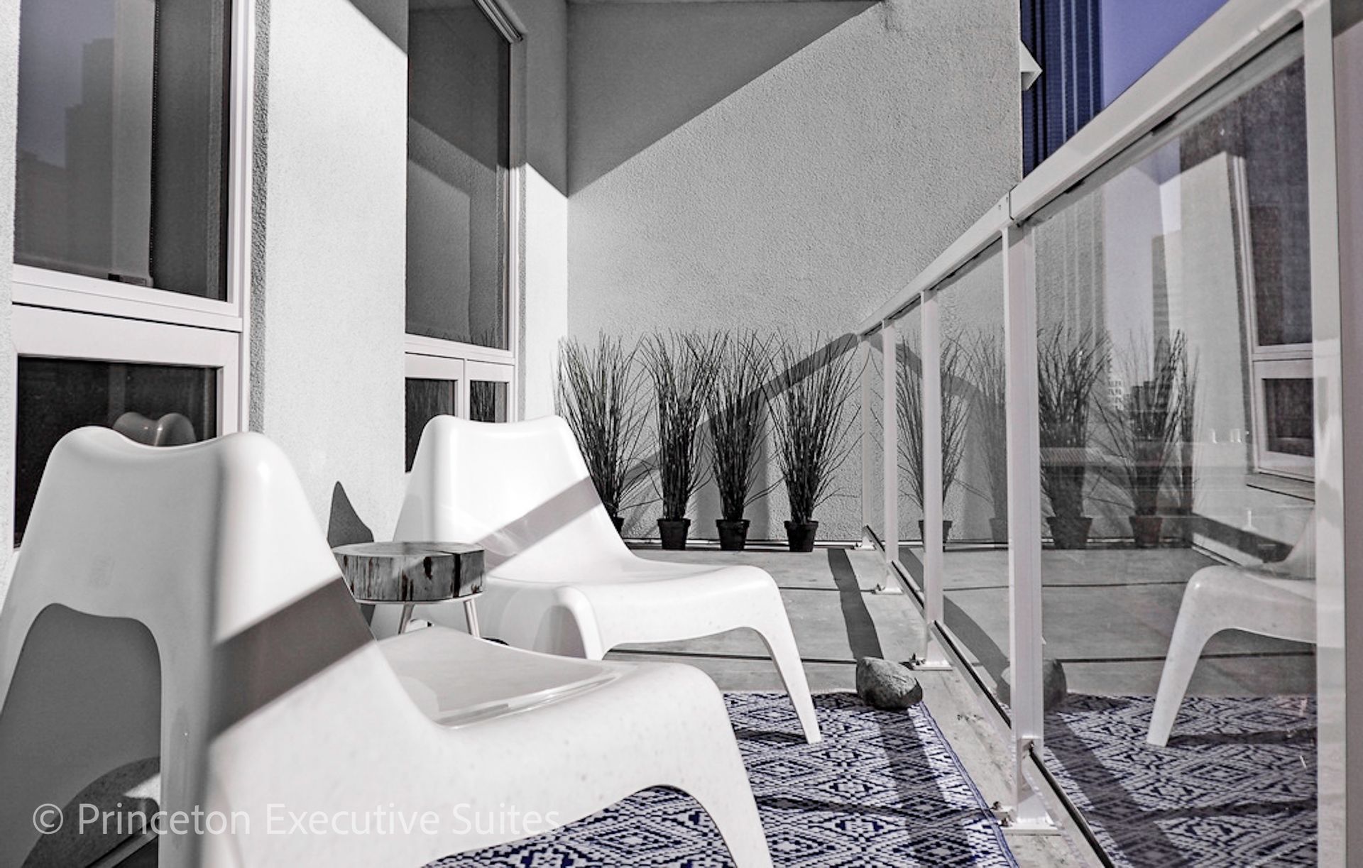 Balcony with two white chairs, small table, and plants, reflecting cityscape.