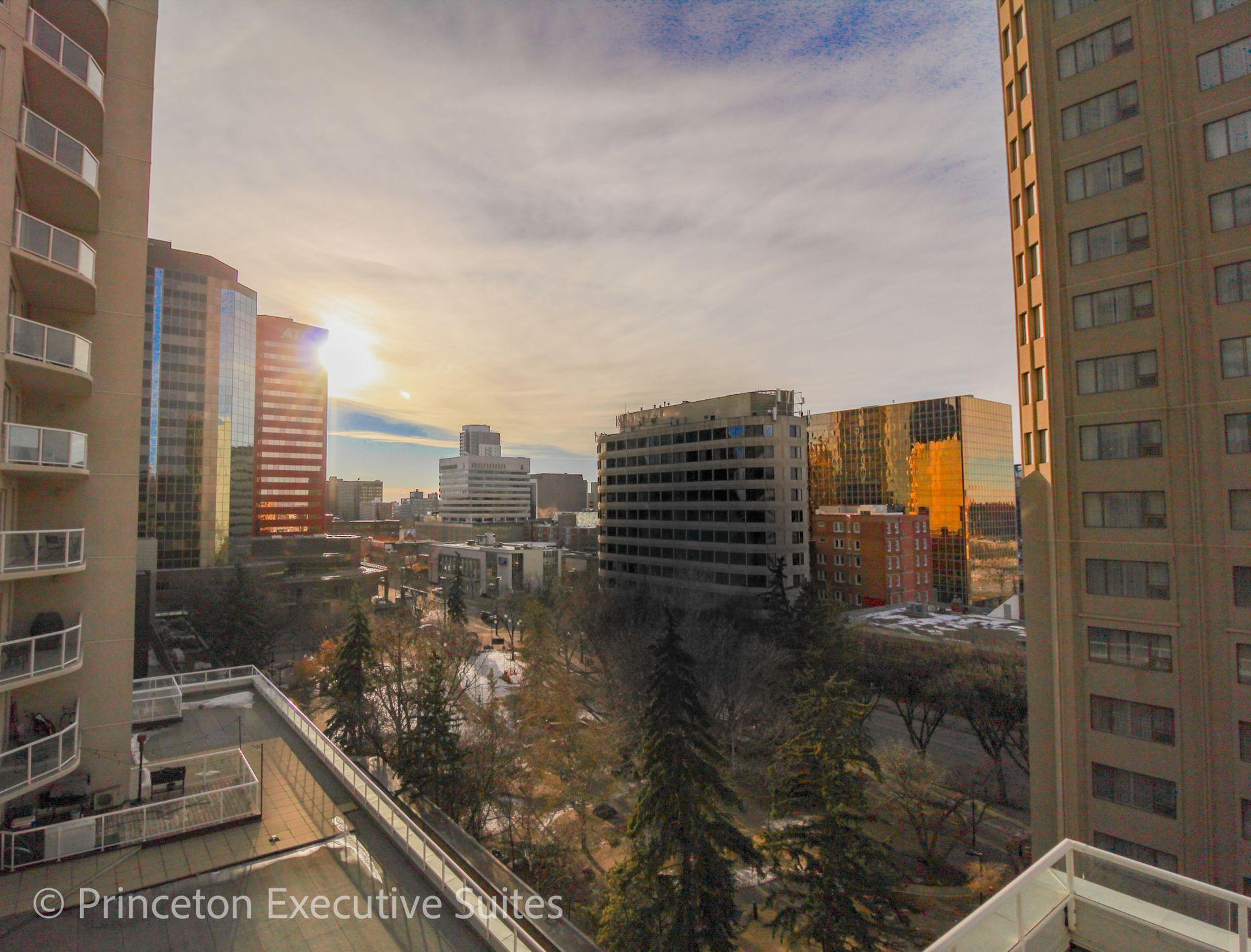 Cityscape with buildings, trees, and a bright sun setting.