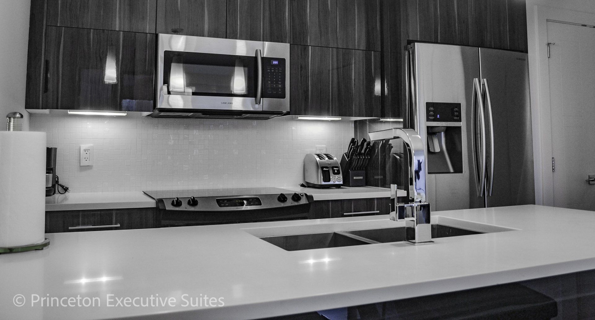 Grey Kitchen with white quartz counter top on counter next to a ceramic cooktop stove and a stainless steel refrigerator