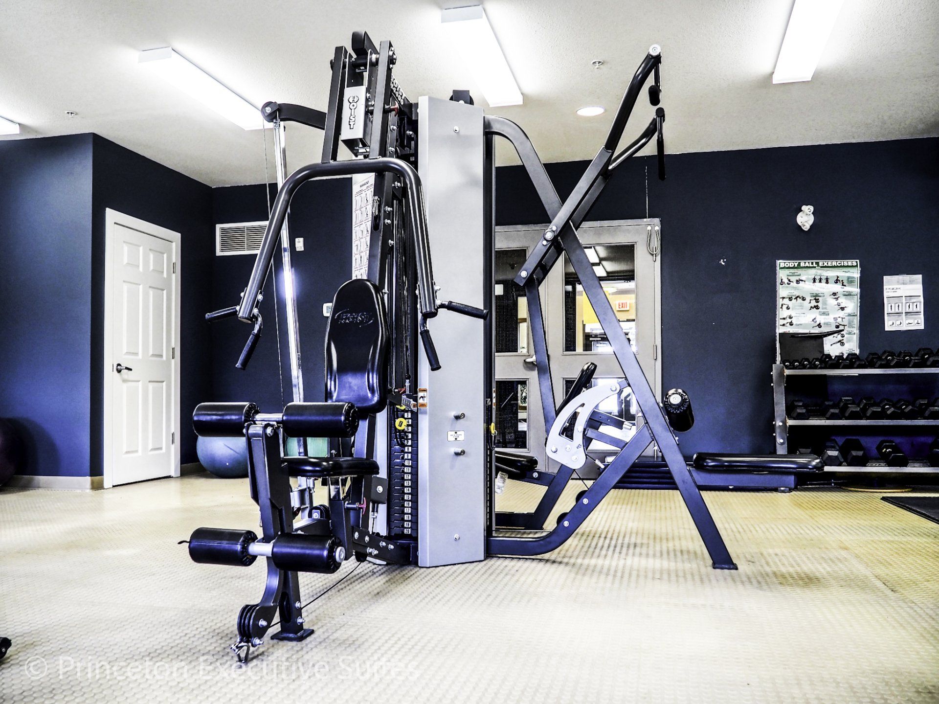 Gym with multi-station weight machine, equipment, and weights against blue walls and light-colored floor. 