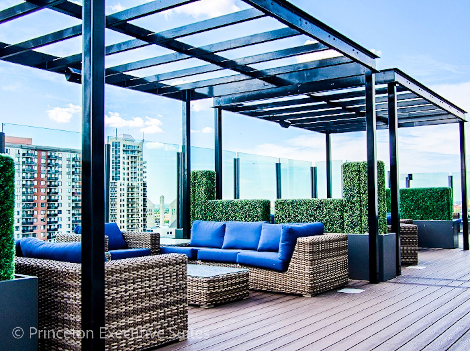 Rooftop lounge with blue sofas, woven chairs, and a pergola. City skyline in background.