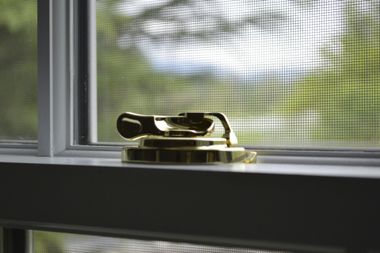 Golden window latch on a white windowsill with a screen window in the background showing green foliage.