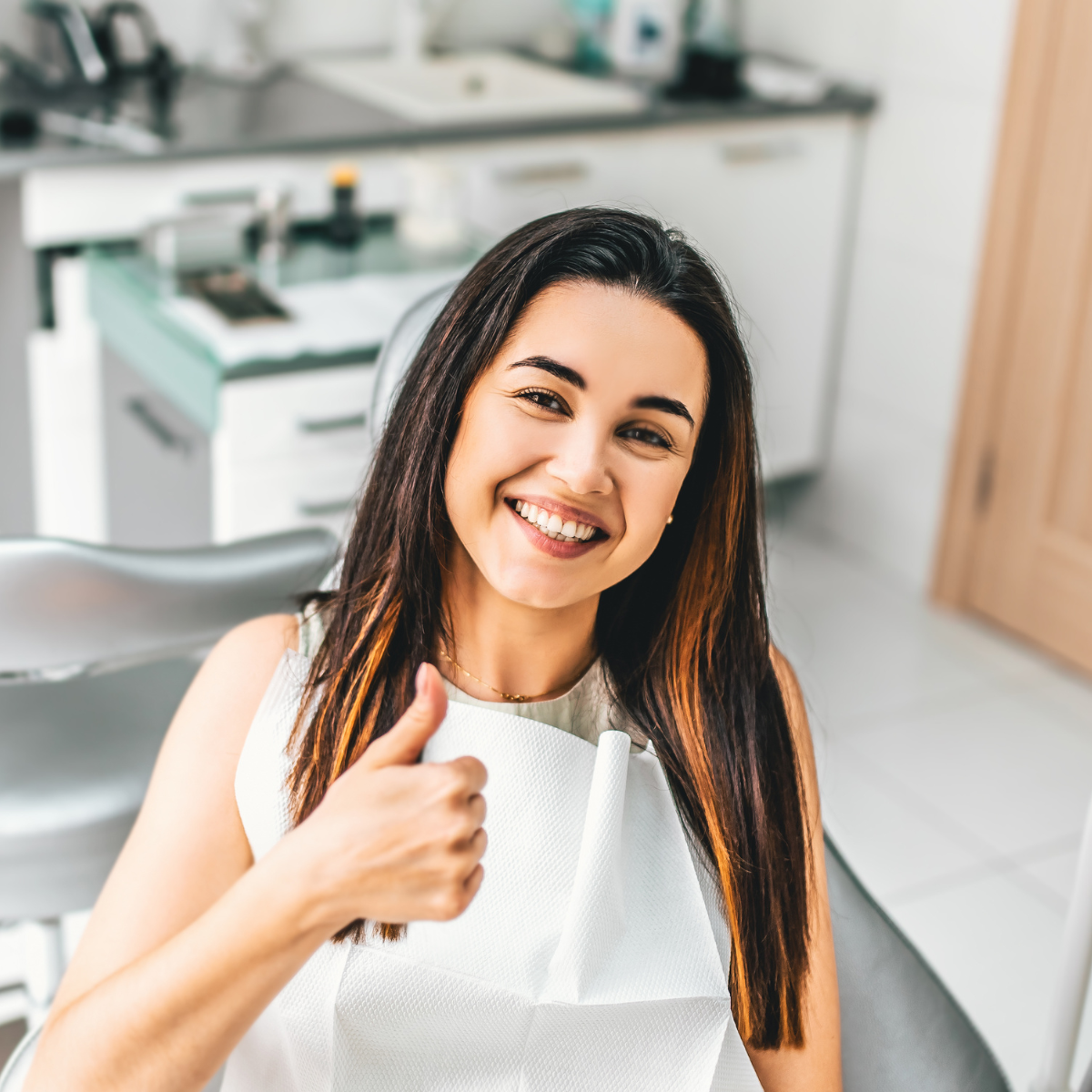 A woman in a dental chair is smiling and giving a thumbs up.