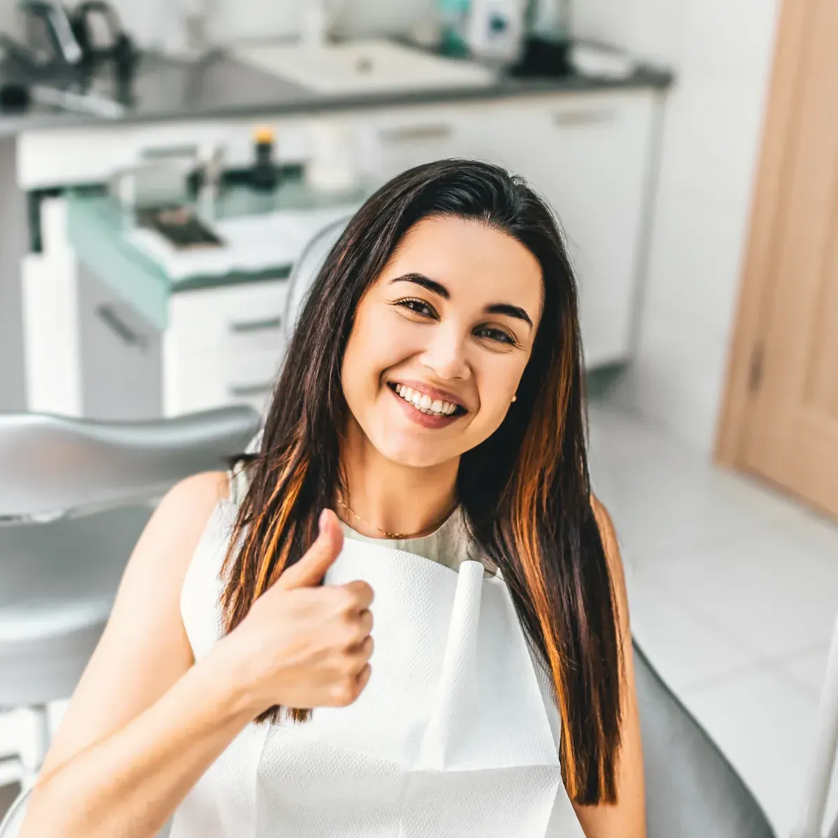 A woman in a dental chair is smiling and giving a thumbs up.