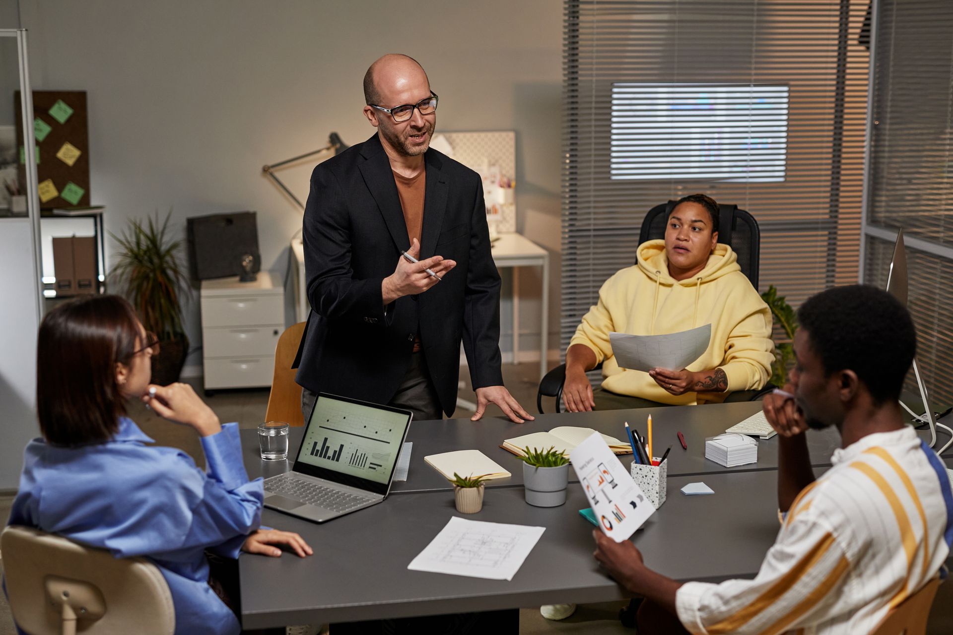 Four people in a dimly lit office at a table. A man stands, gesturing, while others listen and review documents.
