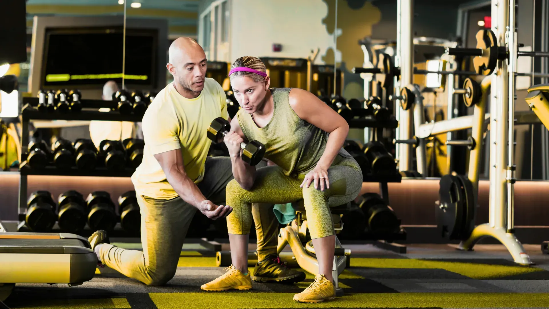 A man is helping a woman lift a dumbbell in a gym.
