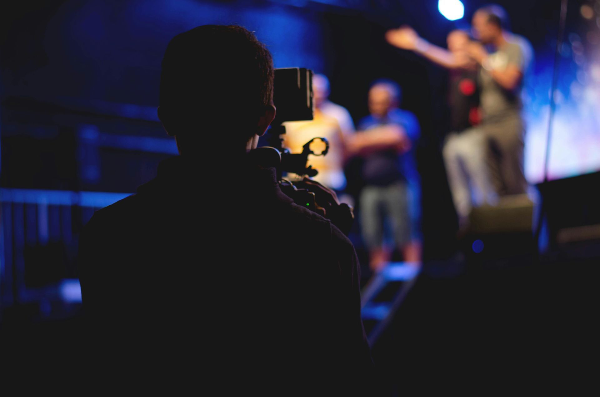 Videographer filming a performer on a stage with others in the background under blue and white lights.