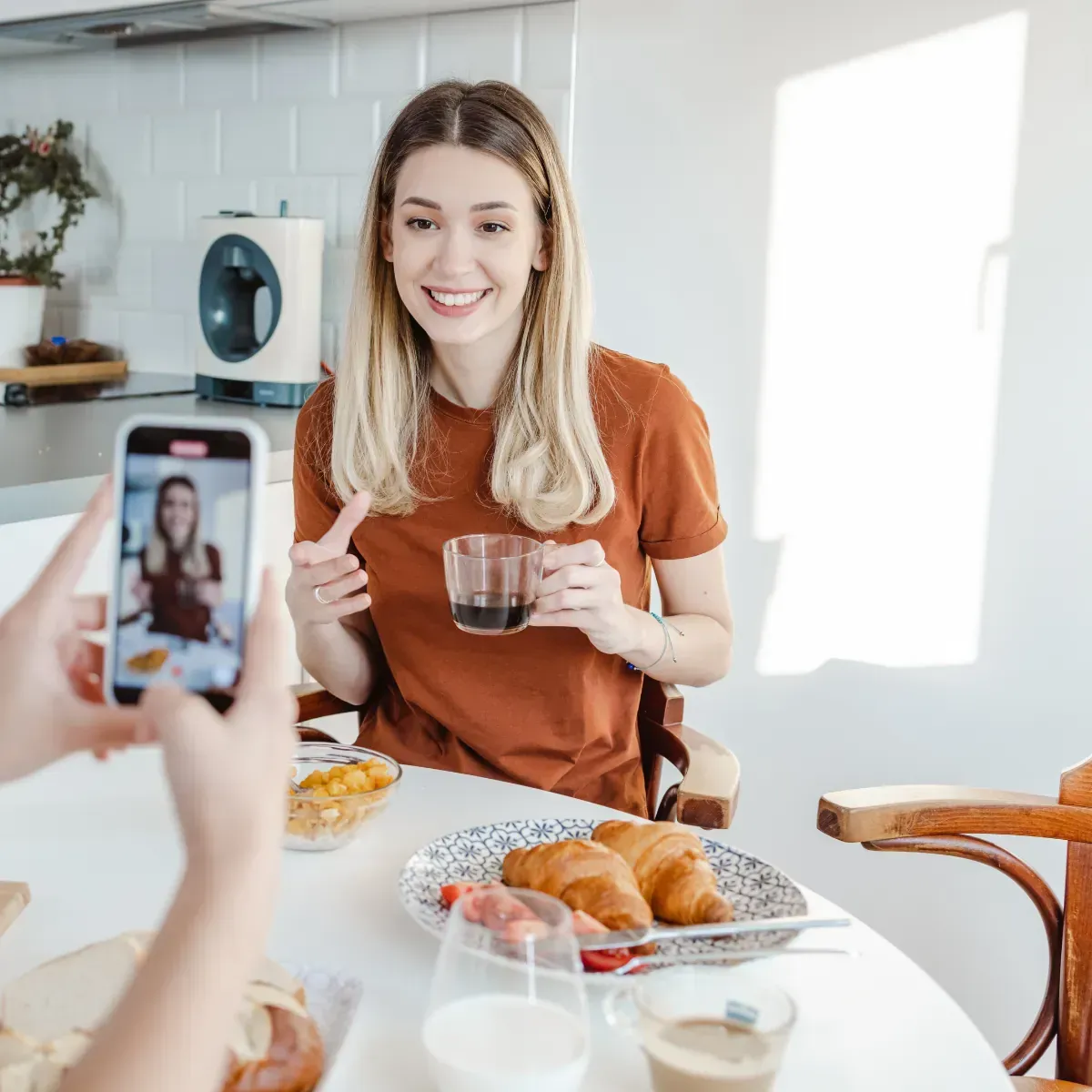 A woman is sitting at a table holding a cup of coffee while another person takes a picture of her.