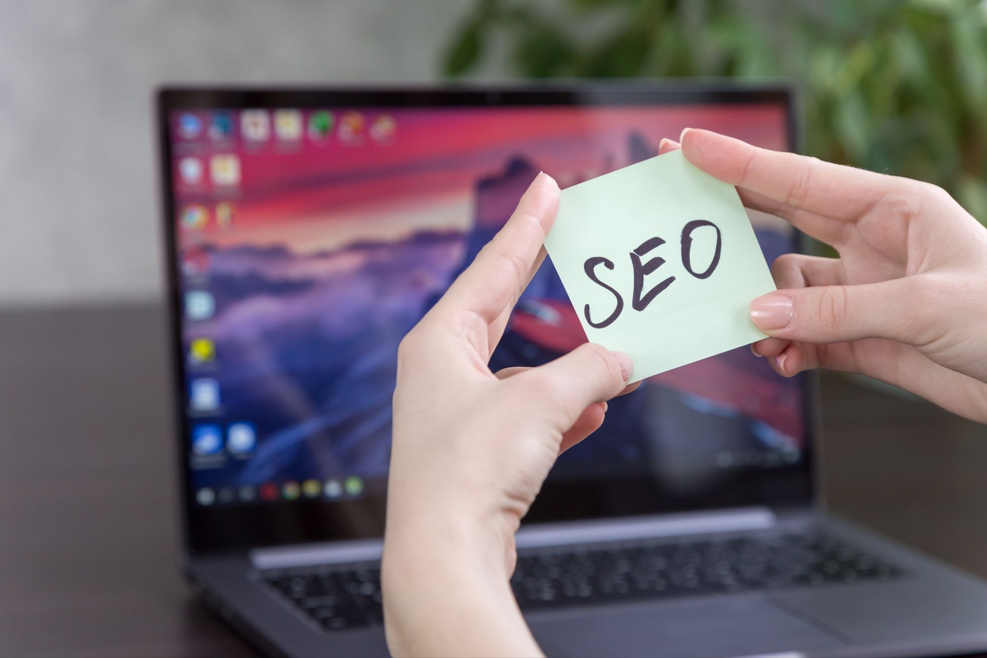 A person is holding a sticky note with the word seo written on it in front of a laptop.