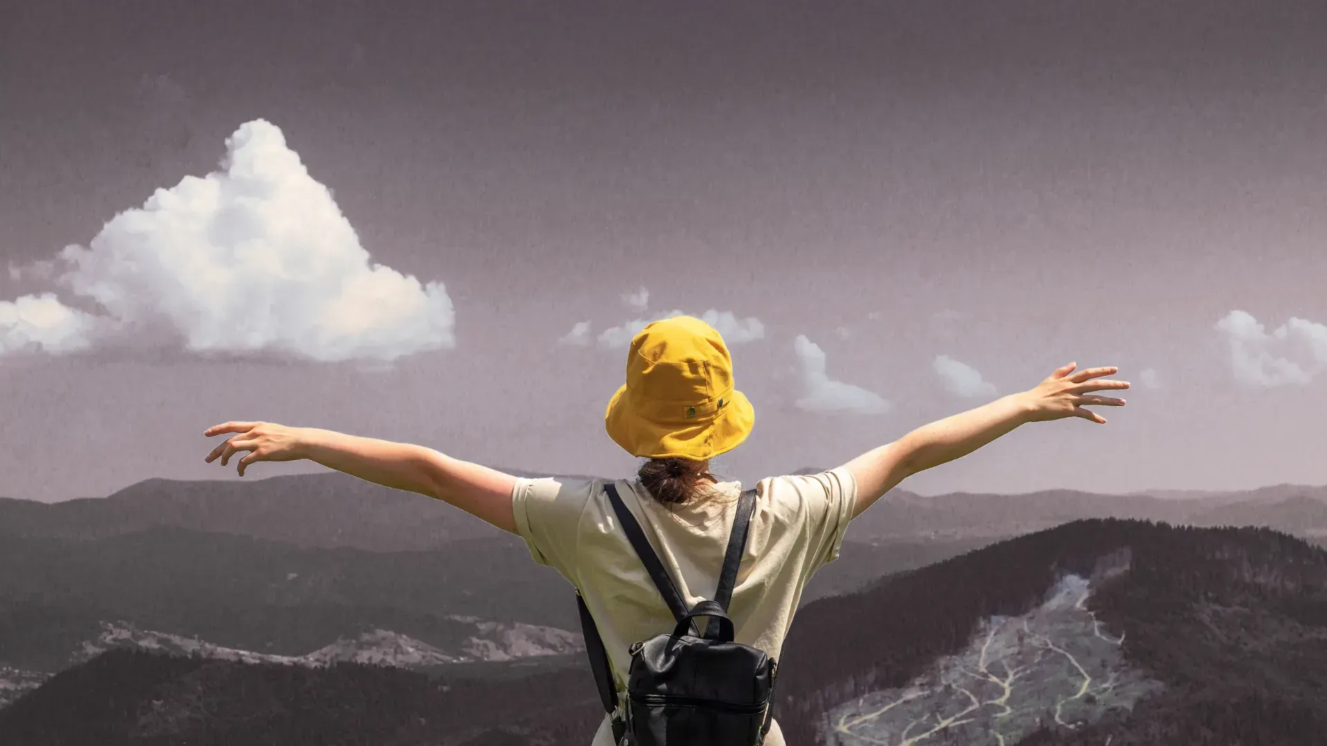 A person wearing a yellow hat and a backpack is standing on top of a mountain with their arms outstretched.