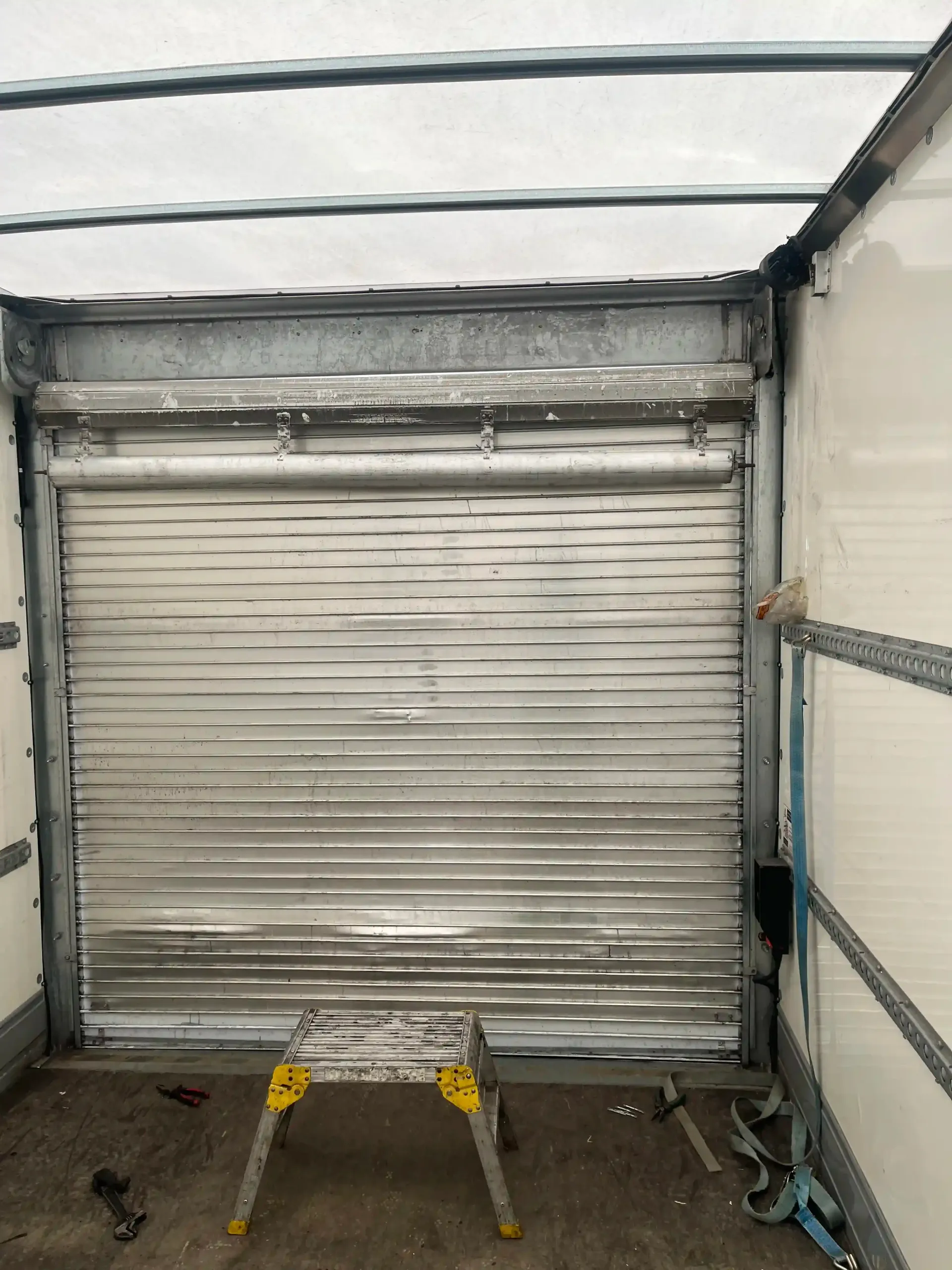 Inside the rear of a box truck, showing a metal roll-up door and a small step stool sitting on the floor.
