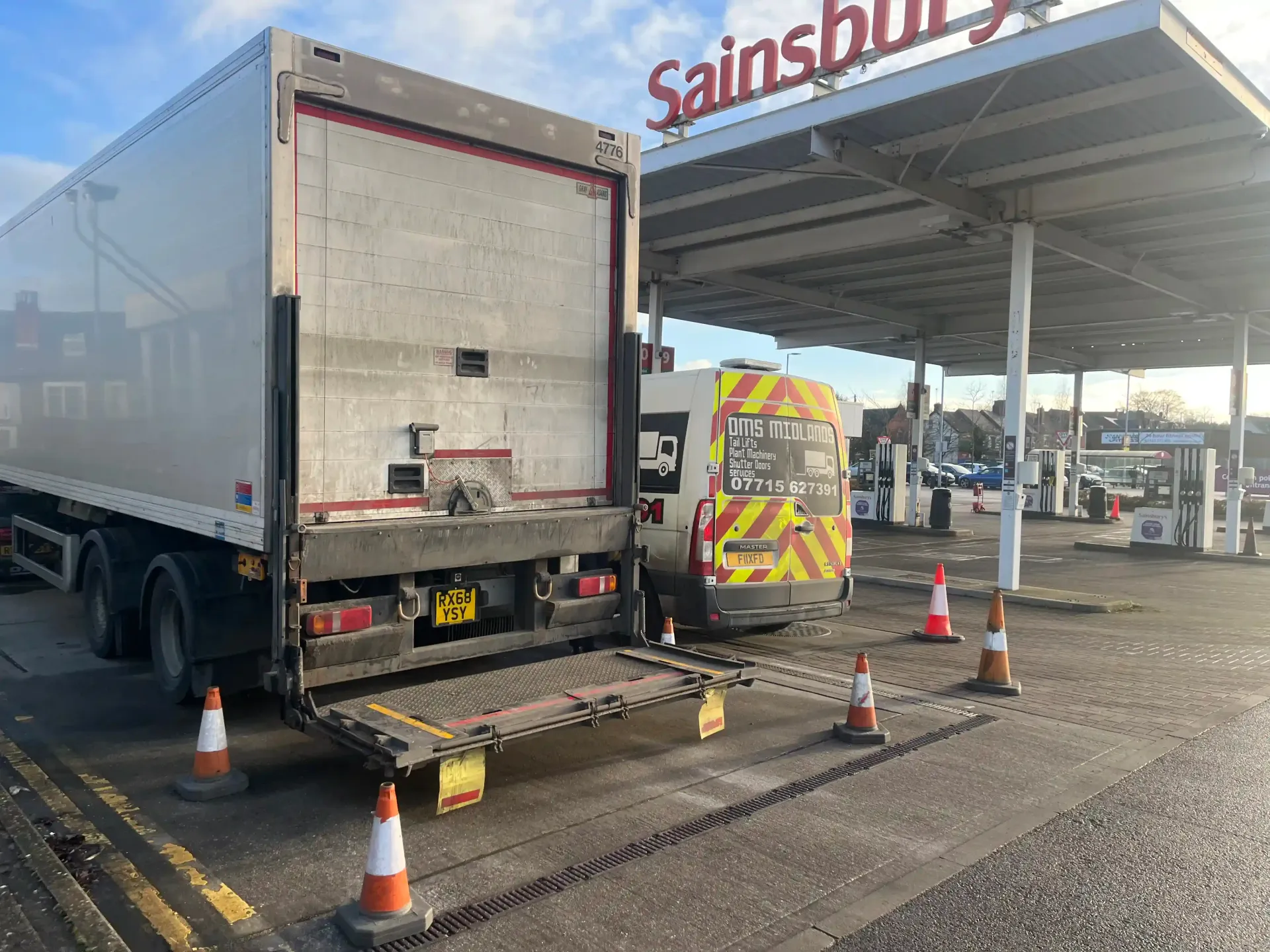 A large white delivery truck with a lowered tail lift parked at a Sainsbury’s petrol station with orange traffic cones.