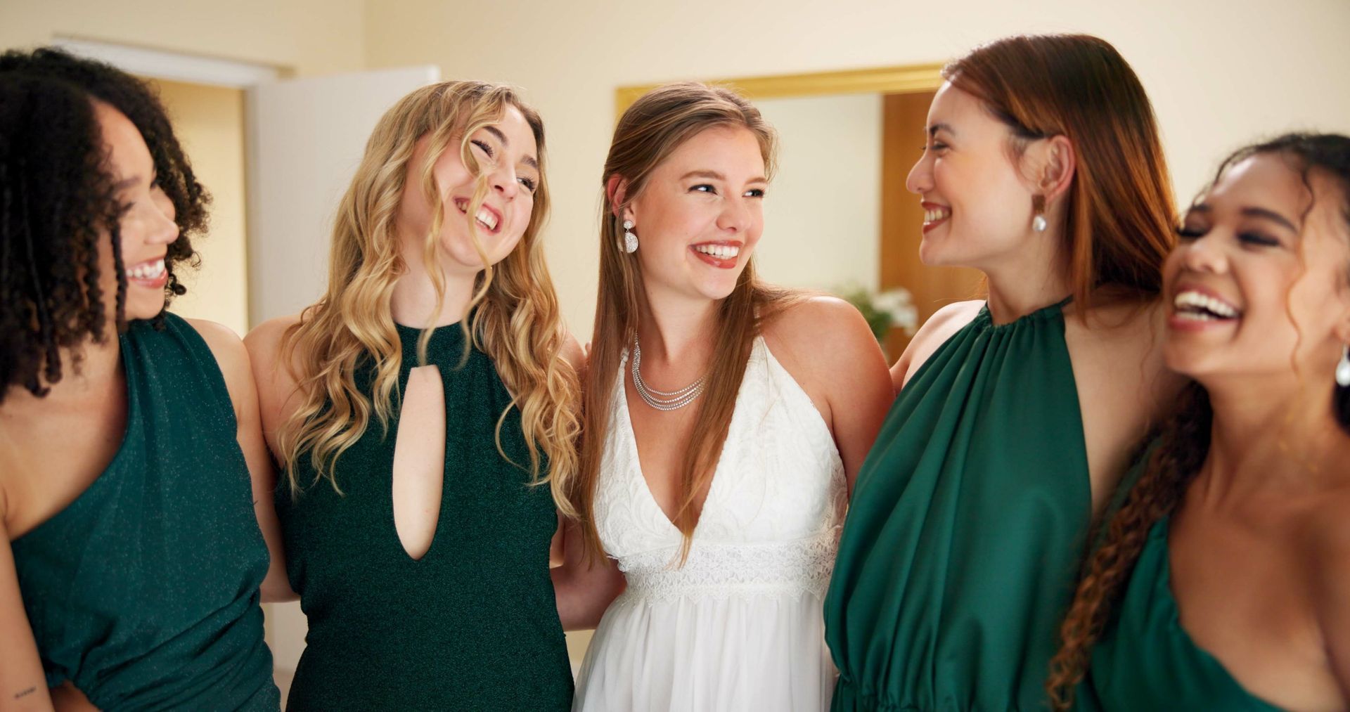 Five young women in gowns laughing. Two in green, one in white, indoors with a mirror.