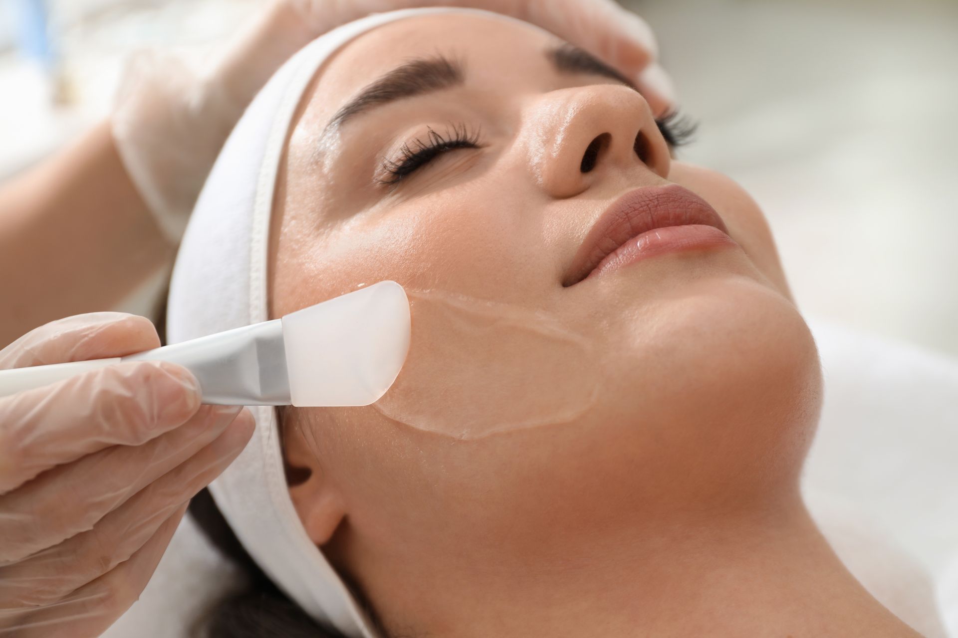 An esthetician in gloves applies a gel treatment to a person's cheek with a brush during a facial spa treatment.