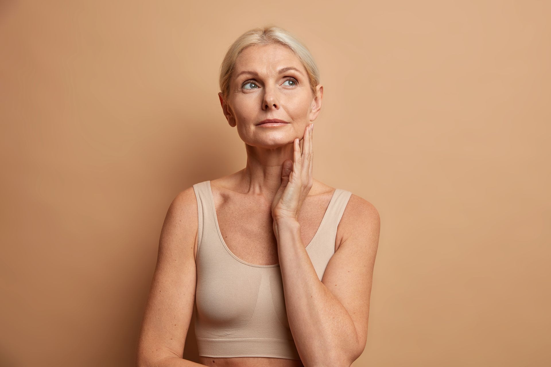 A person in a beige top looking upward with a thoughtful expression, hand touching their chin against a tan background.