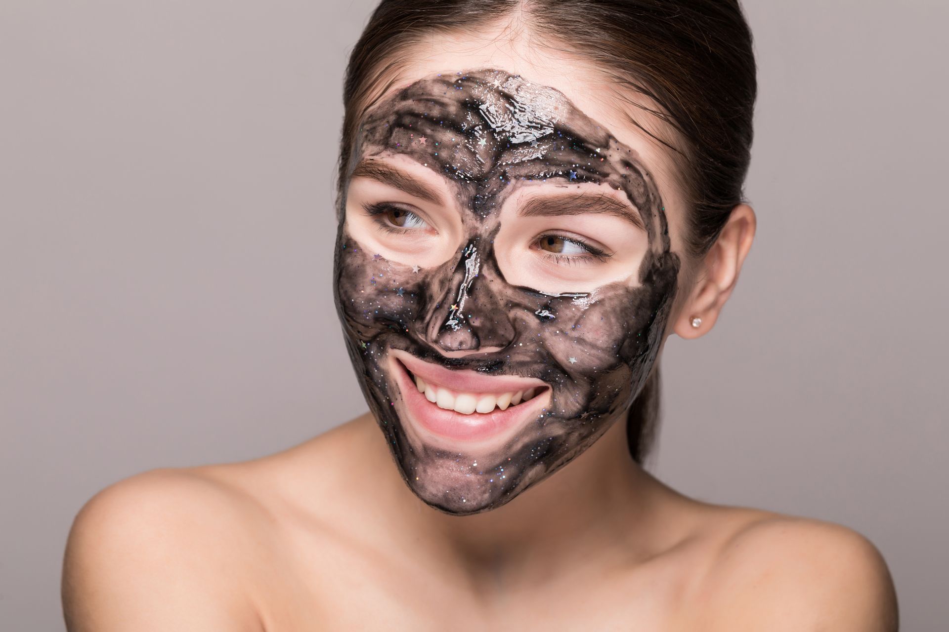 A woman is wearing a charcoal mask on her face and smiling.