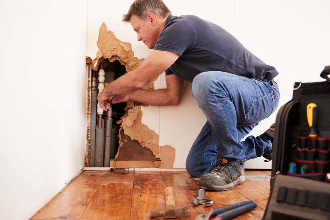 Repair worker kneeling and cutting into a wall beside exposed pipes in a damaged room