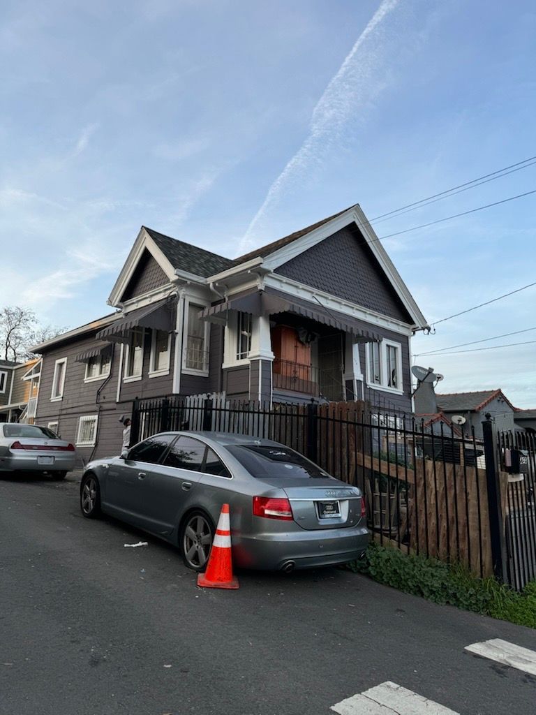 Gray house with silver car parked on an inclined street; orange traffic cone sits nearby.