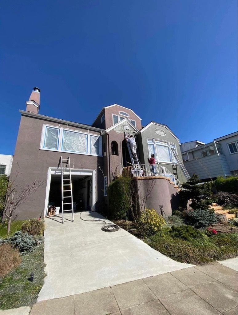 House exterior being painted, with ladders and painters visible. Dusty rose stucco with blue sky.