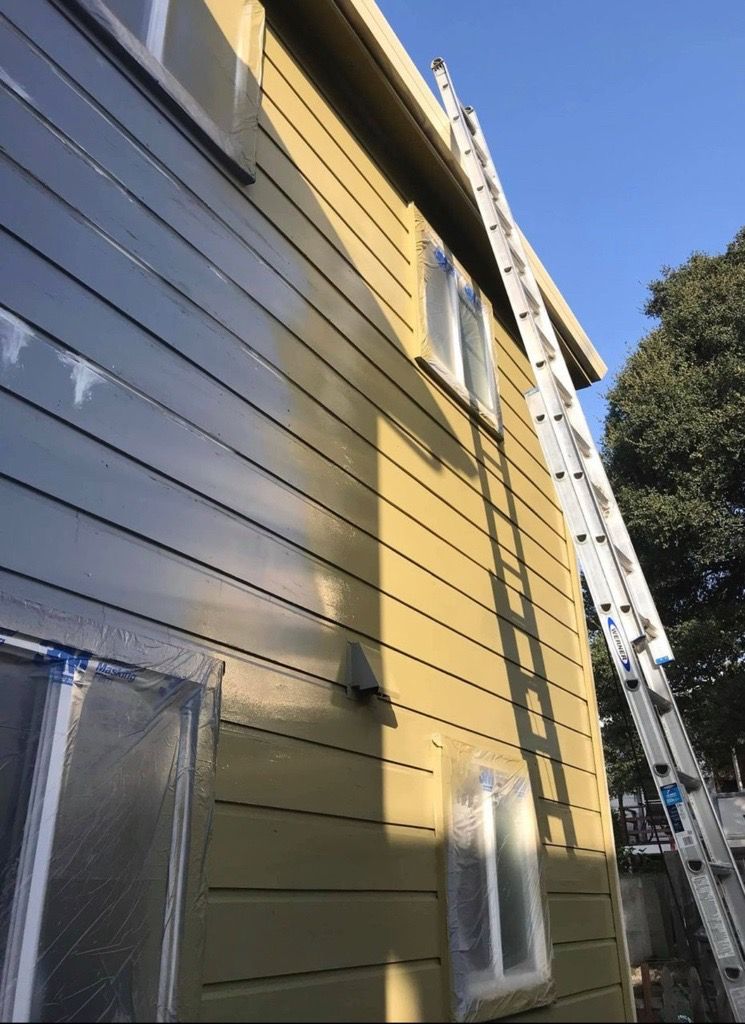 Side of a house being painted, with a tall ladder extending to the roof. Windows are taped, exterior is yellow-green.