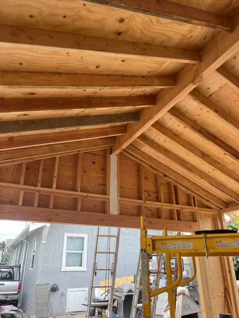 Wooden roof construction with exposed beams, house, ladder, and scaffolding in the background.