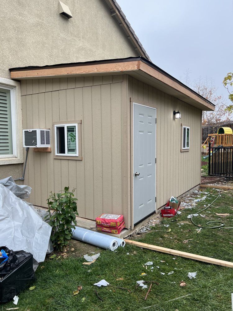 Tan shed with a white door, windows, and an AC unit attached to a beige house, surrounded by grass.