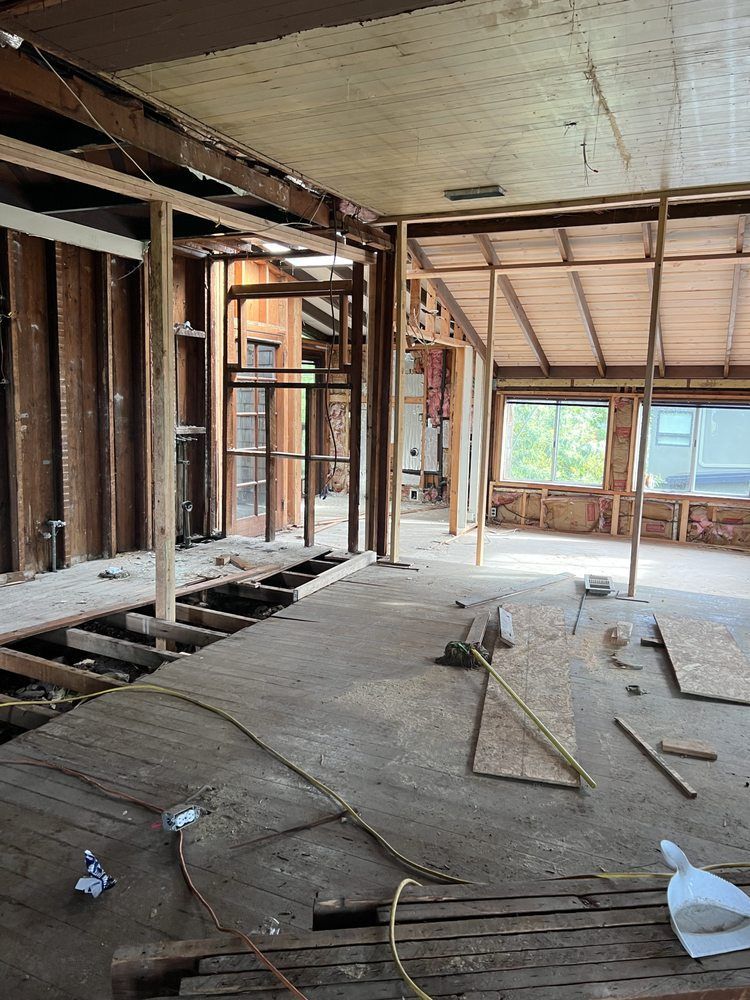 Interior of a house under construction; exposed wooden framing, bare concrete floor, and natural light from windows.