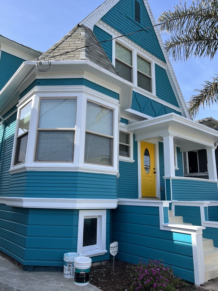 Teal house with white trim and a yellow door; gray roof; two paint cans on the ground. Teal house with white trim and a yellow door; gray roof; two paint cans on the ground.