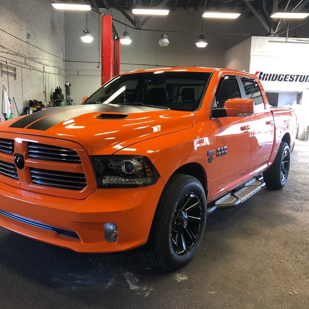 Bright orange pickup truck in a garage, angled front view with black wheels and hood stripes