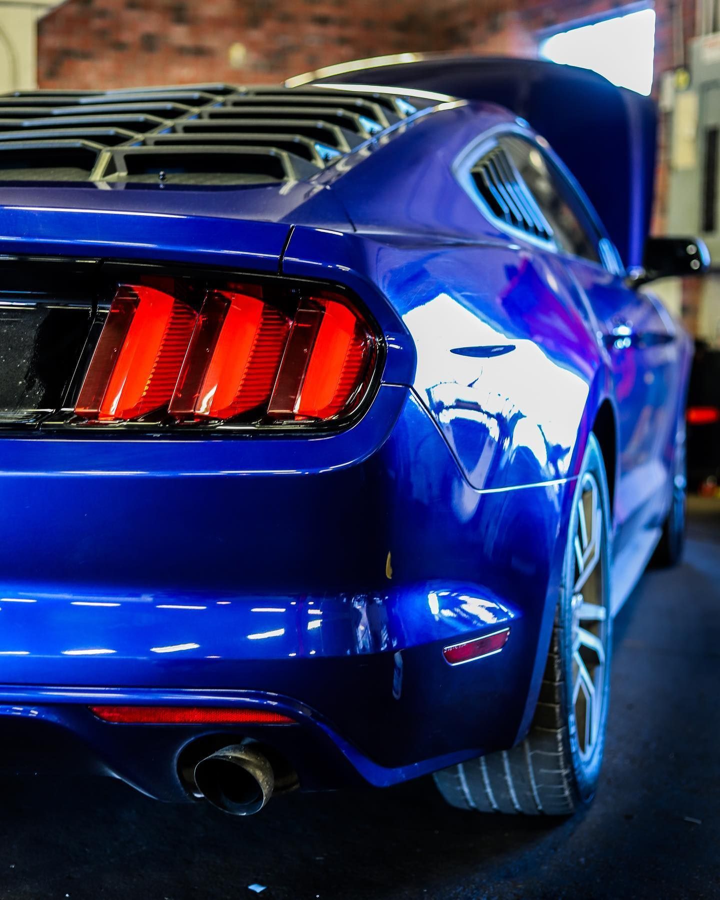 Blue sports car rear view with red taillights in a garage, showing a sleek spoiler and chrome wheel.