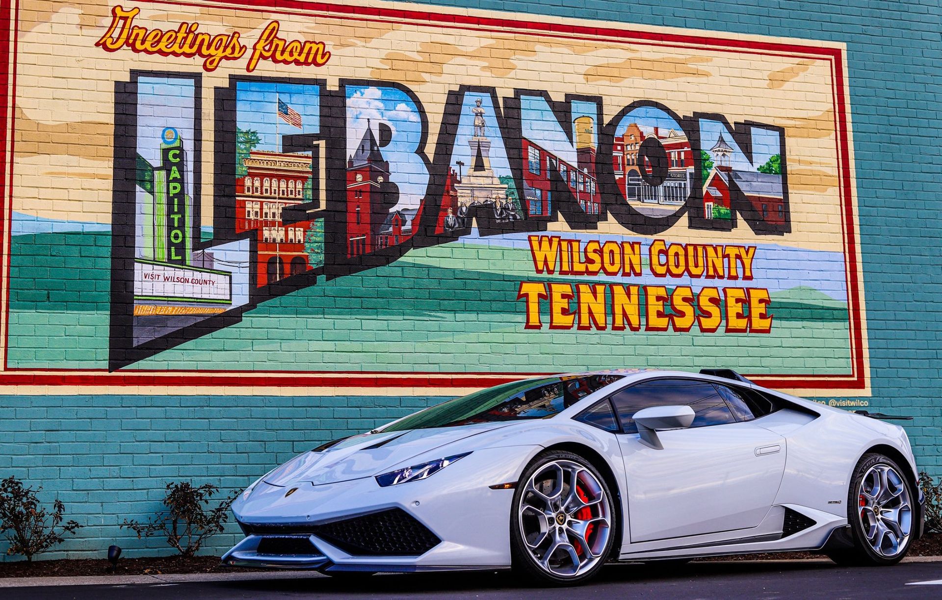 White sports car parked beside a colorful mural reading “Bacon, Wilson County, Tennessee.”