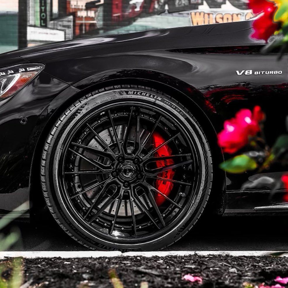 Close-up of a black sports car’s front wheel and fender, with red brake calipers and pink flowers in the foreground