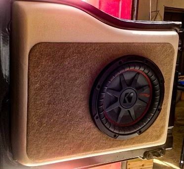 Beige speaker enclosure with a round black-and-red subwoofer in a car audio setup.