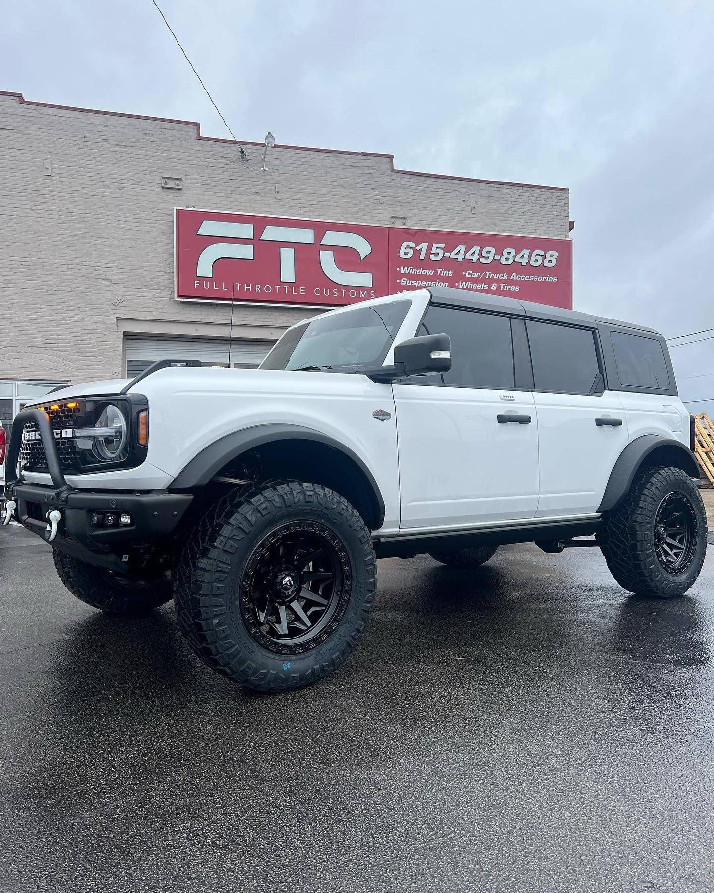 White lifted Ford Bronco parked on wet pavement outside a building with an FTC sign