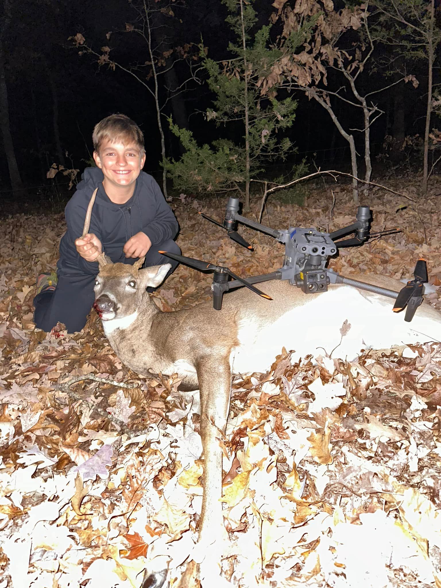 Two men pose with a harvested deer. They're sitting in tall grass, one with a rifle, both smiling, under a dark sky.