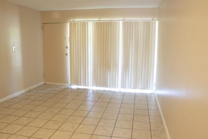 Empty beige living room with tiled floor and vertical blinds over a sliding glass door