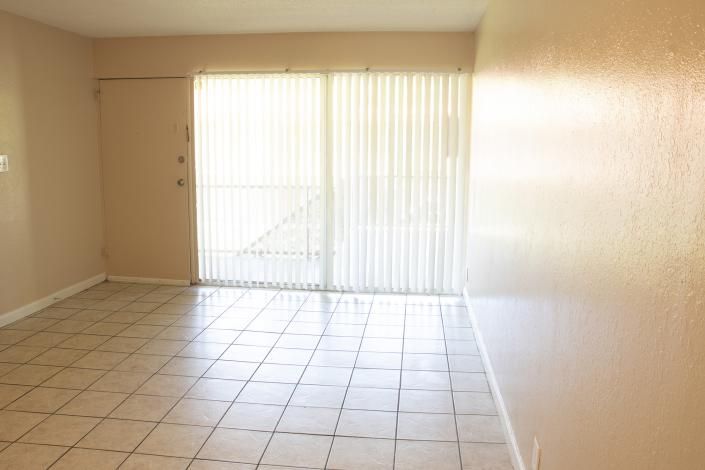 Empty beige living room with tiled floor and sliding glass door with vertical blinds.
