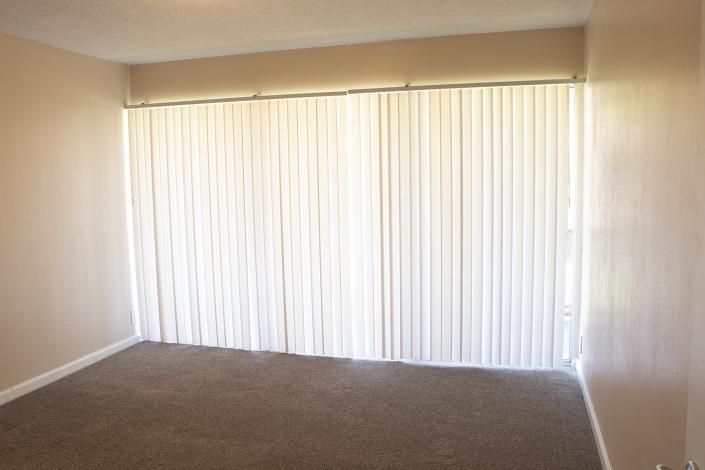 Empty carpeted room with beige walls and white vertical blinds covering a large window.