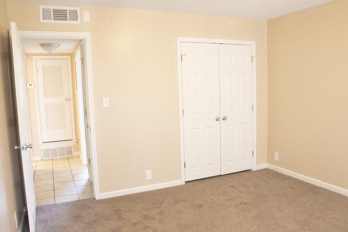 Empty beige bedroom with carpet, white closet doors, and a hallway view to tiled area