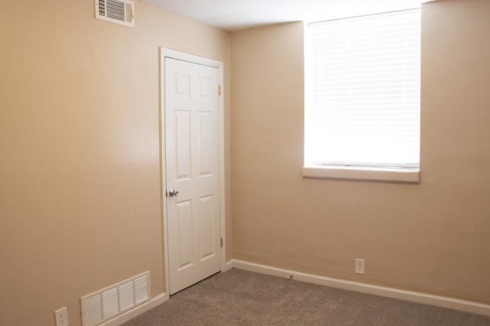 Empty beige bedroom with white door, window blinds, and carpeted floor.
