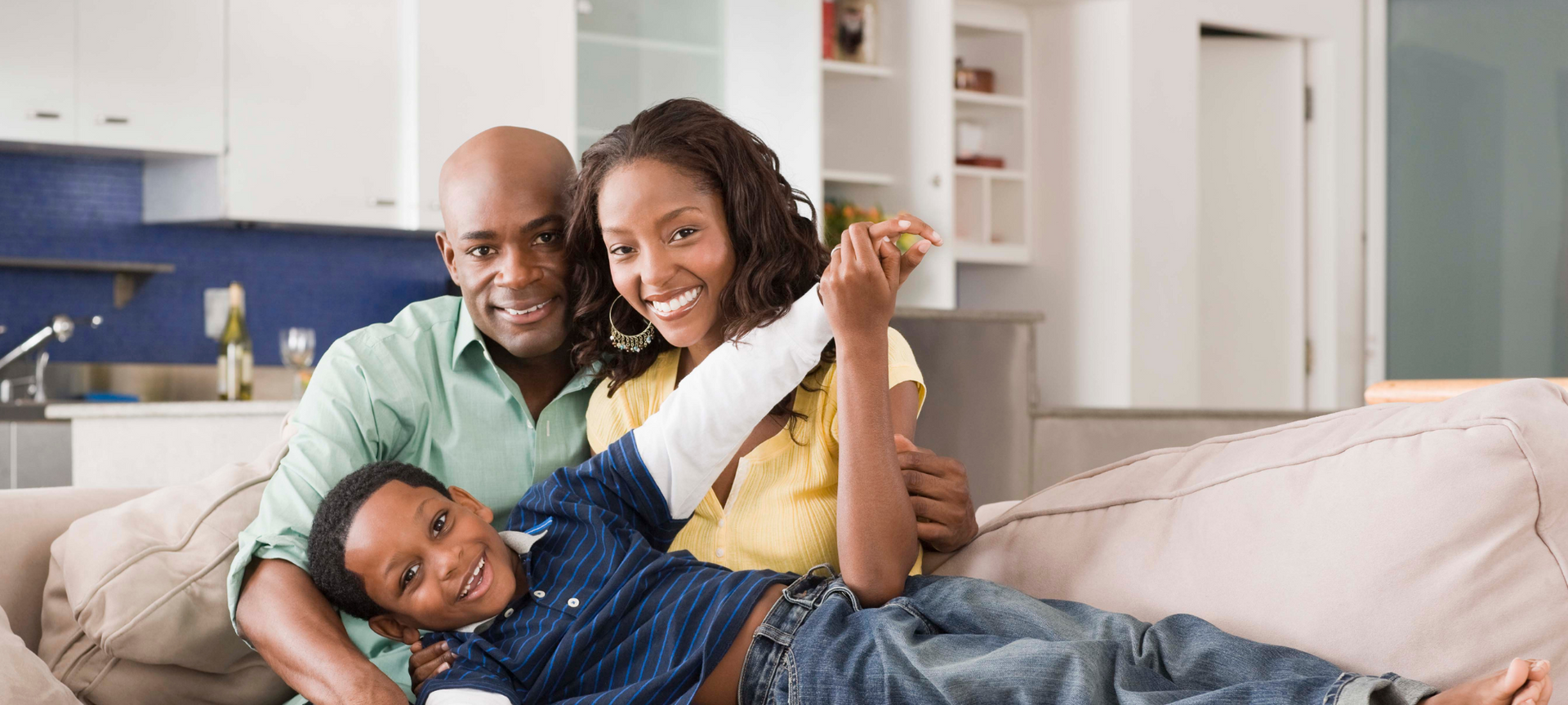 Family smiling on a couch in a bright living room, with a child lying between two adults