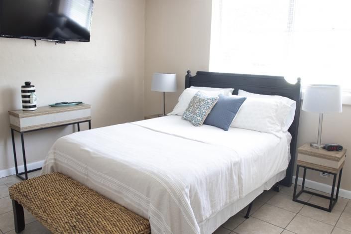 Bright bedroom with a white bed, dark headboard, wall-mounted TV, and bedside tables