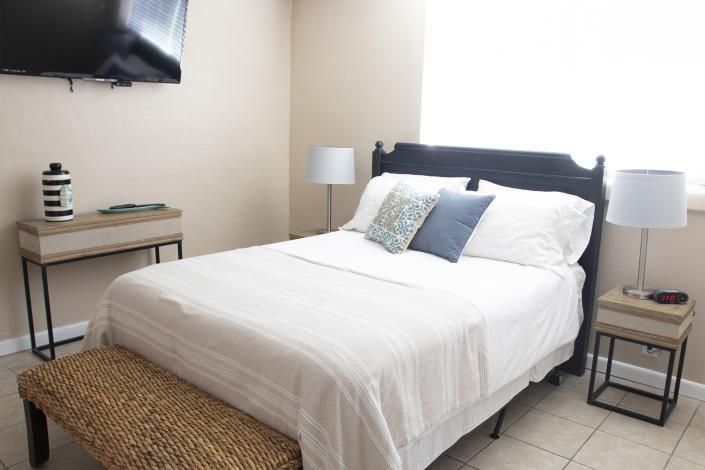 Bright bedroom with a white bed, dark headboard, bedside lamp, TV, and woven bench at the foot.