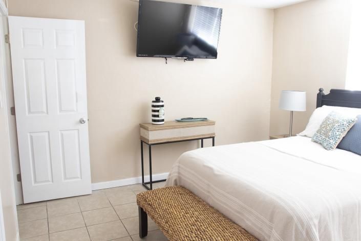 Bright bedroom with white bed, wall-mounted TV, desk, lamp, and tiled floor
